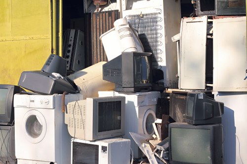 A large pile of discarded household appliances and electronic waste situated outdoors, consisting of washing machines, microwave ovens, old television sets, computer monitors, and various metal and plastic components. The appliances are stacked and haphazardly arranged on a concrete surface in front of a metal storage shed, with some items leaning against each other. The washing machines are predominantly white, with visible control panels and circular doors. The older televisions and monitors display dark screens, and some are partially covered with dirt or dust. Surrounding the pile, there are a few loose wires and smaller debris, indicating recent clearance activity. The background includes a partially open door of the shed and a worn, brown metal wall. The environment suggests an informal outdoor rubbish collection area, with evidence of electronic waste typical of house clearance services such as Elephant and Castle House Clearance operating within the London borough. The overall scene captures the variety of bulky, outdated household items awaiting removal and disposal, emblematic of a typical rubbish clearance project in the South London area postcode area.
