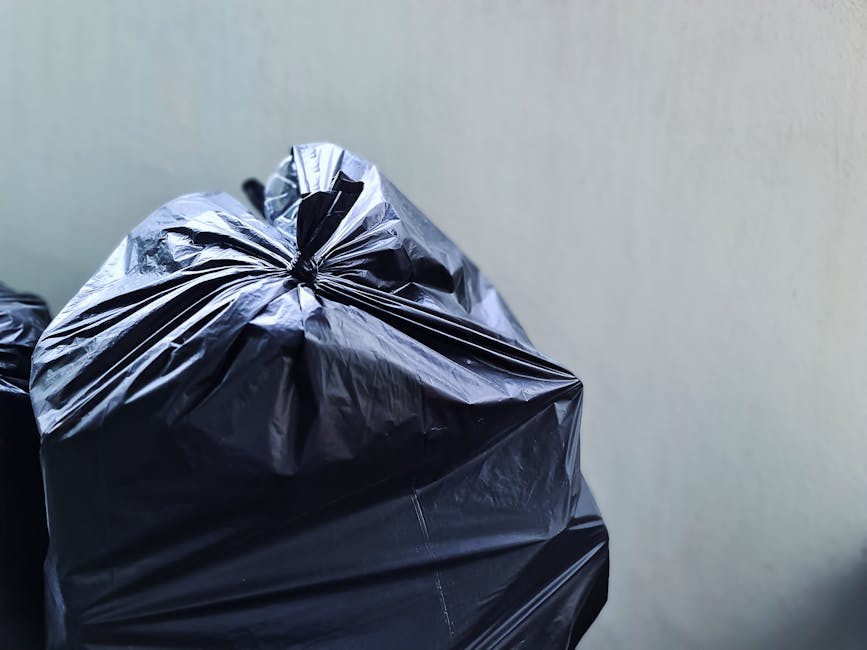 Close-up of several black plastic rubbish bags filled with waste, tightly knotted at the top, placed against a plain light grey wall background. The bags are made of glossy, crinkled plastic material,