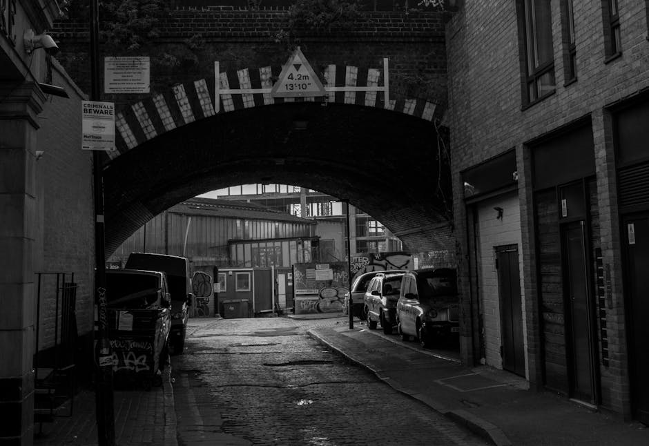A black-and-white photograph of a narrow urban alleyway beneath a brick archway, with several parked vehicles including a dark van on the left and multiple small cars on the right side. The alley surf