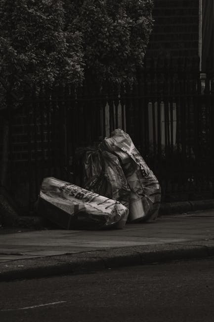Two large black plastic garbage bags filled with waste are positioned on the curbside pavement next to a dark metal fence. The bags appear to be tightly knotted and contain household or garden refuse,