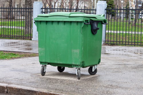 A medium-sized green plastic wheelie bin with a closed lid, positioned on a paved sidewalk in front of a black metal fence with vertical bars and concrete posts. The bin has four small wheels at its base, enabling mobility, and features a black handle on the front right side. The background includes a grassy area with leafless trees and a cloudy sky, suggesting a residential or park setting in an urban environment, possibly in or near Elephant and Castle. The surface appears damp, and the scene is indicative of municipal rubbish collection services, as handled by companies like Elephant and Castle House Clearance, operating within the local boroughs.
