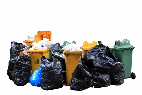 A collection of various waste bins and trash bags positioned outdoors on a paved surface, with some bins in yellow, green, and orange colors, and a number of black and blue plastic rubbish bags piled around them. The bins have closed lids, with a few lids partially open, revealing waste inside. The scene appears to be part of waste collection or rubbish removal activity, set against a plain white background with no surrounding environment visible. The diversity of container types and the arrangement suggest a typical rubbish disposal area, possibly associated with domestic or commercial clearance services in London. The image emphasizes the volume of waste accumulated and is relevant to waste management and rubbish removal services offered by Elephant and Castle House Clearance, illustrating the typical contents and setup for rubbish clearance tasks in the area.