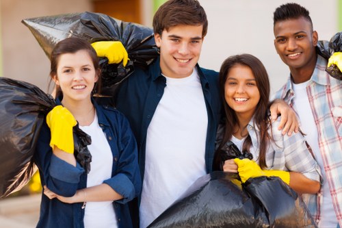 A group of four young adults, two women and two men, standing outdoors on a paved residential sidewalk, each holding large black rubbish bags filled with waste. The individuals are smiling and appear to be in good spirits, wearing casual clothing with some of them dressed in jackets and others in shirts, and all have yellow cleaning or disposal gloves on their hands. Behind them, there is a blurred background of a house or building exterior with neutral tones. The scene suggests participation in a community rubbish clearance activity, aligning with services offered by Elephant and Castle House Clearance. The image visually emphasizes teamwork and environmental responsibility within a London suburb setting, highlighting a friendly and organized approach to rubbish removal for local homes and properties within the postcode area near Elephant and Castle.
